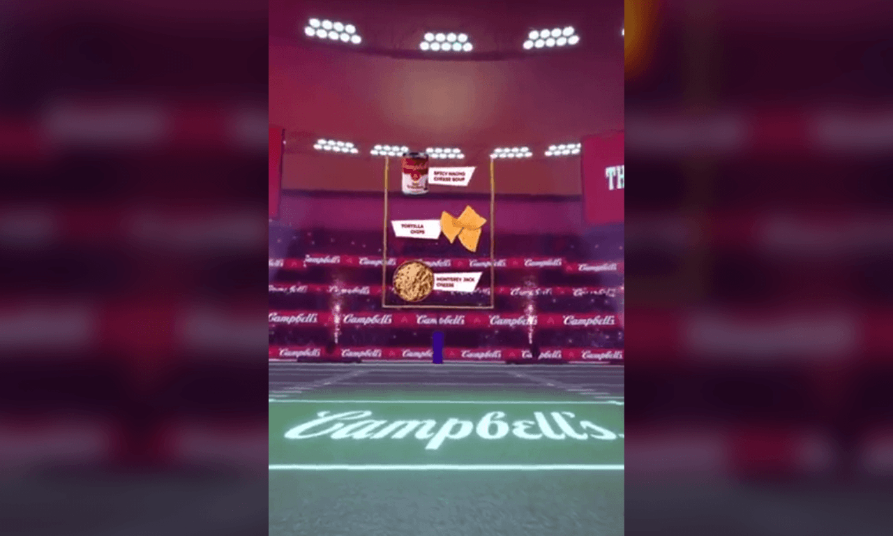 A 3D animated football stadium branded with Campbell's, featuring food items like soup, tortilla chips, and cheese stacked vertically between yellow goalposts.