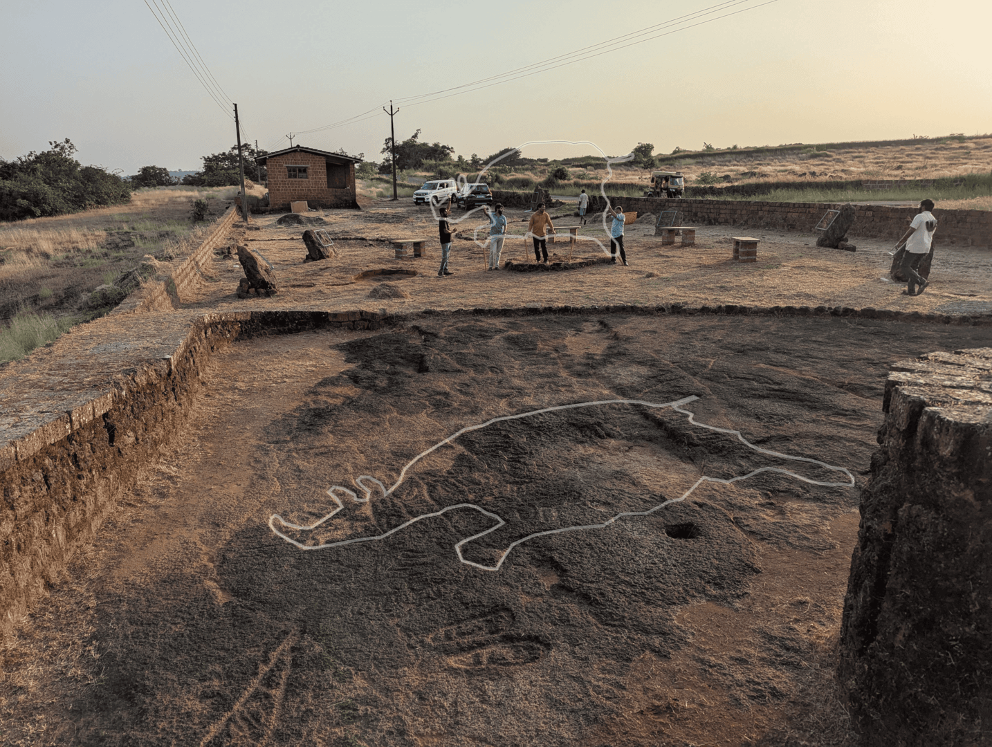 Wide view of an outdoor archaeological site of chave dewood with a digital white outline tracing a large animal petroglyph on a flat rock surface in the foreground, while a research team works in the background.