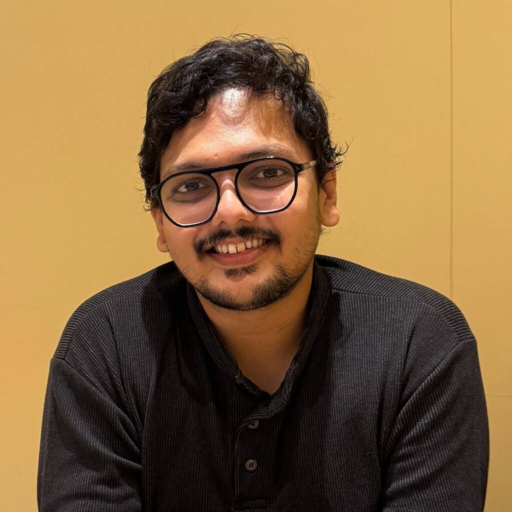 A headshot portrait of a man with dark curly hair, glasses, and a mustache, wearing a black shirt against a warm yellow backdrop.